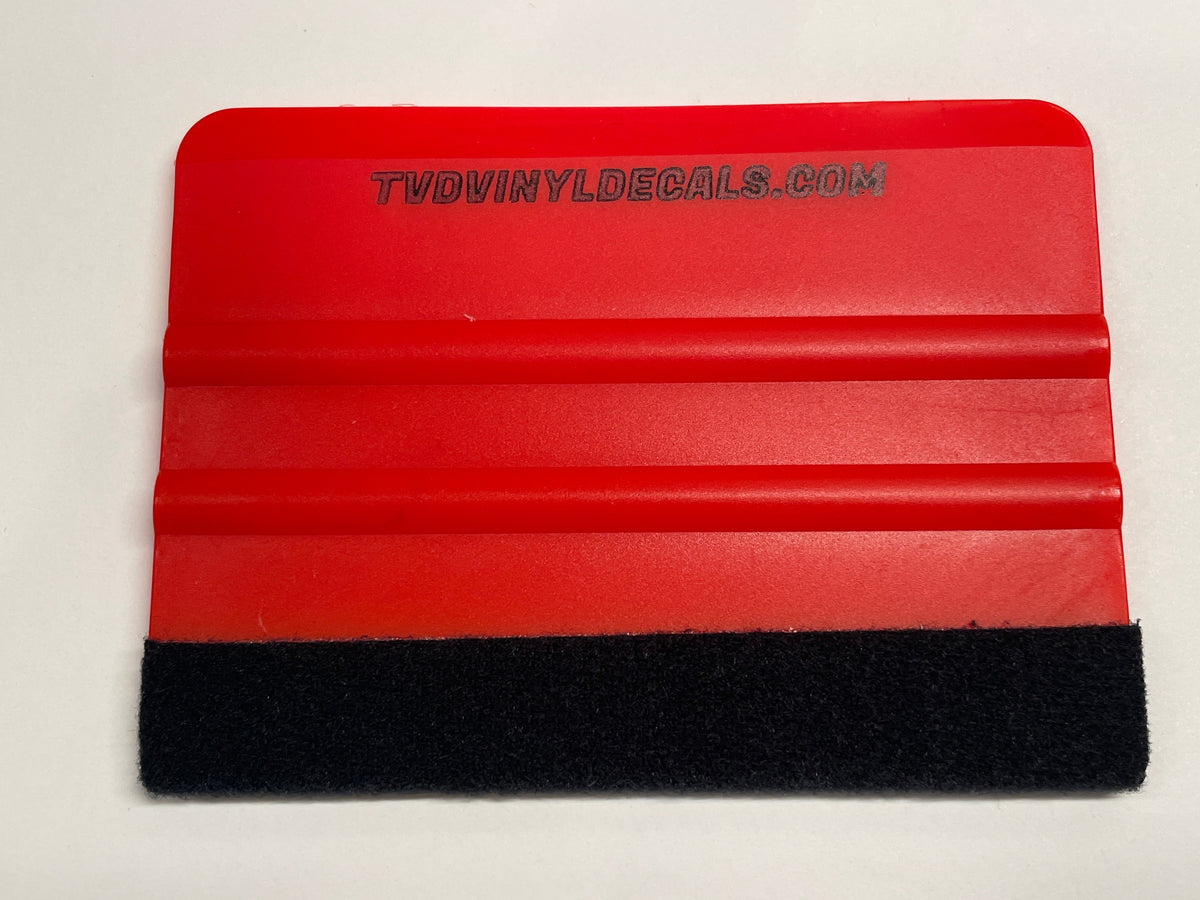 A red plastic squeegee tool with a black  felt strip on one  side and horizontal ridges, featuring the text 'TVDVINYLDECALS.COM' printed on the red surface, shown on a plain off-white background.